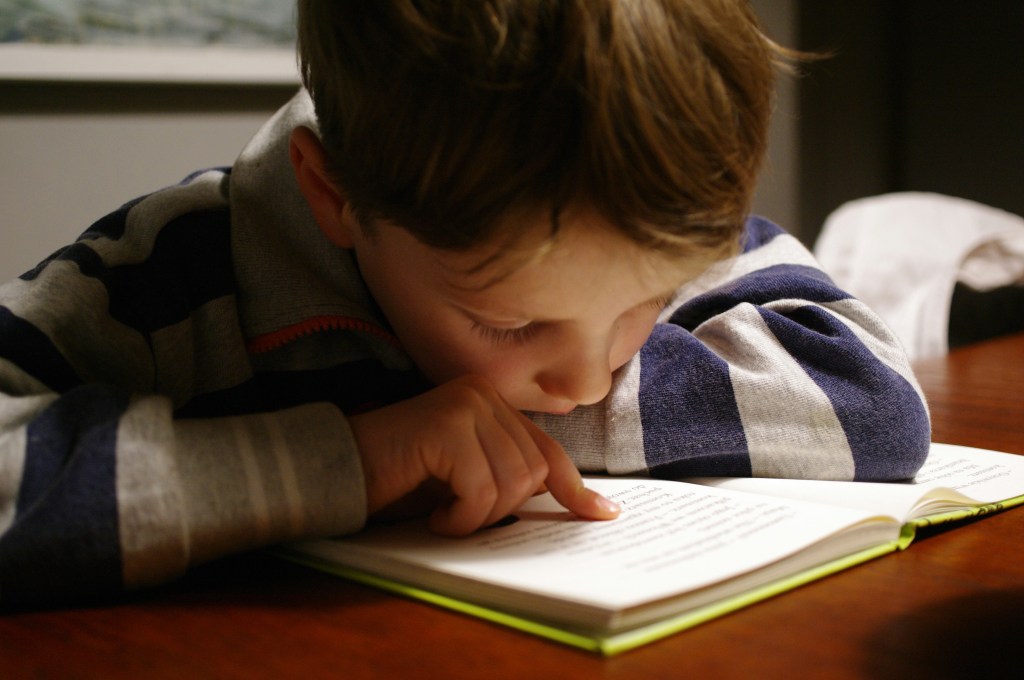 A young boy reading a book intently, resting his chin on his arms as he follows the text with his finger.