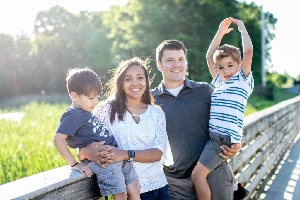 A happy family of four on a wooden bridge, with a mother holding one son and a father lifting another son in the air, surrounded by green grass and trees under sunlight.